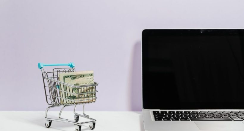 macbook pro on white table beside a miniature shopping cart with money