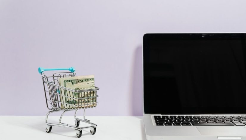 macbook pro on white table beside a miniature shopping cart with money