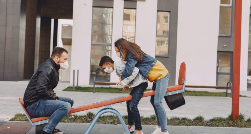 family in mask swinging on seesaw on playground