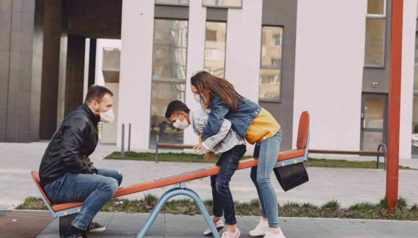 family in mask swinging on seesaw on playground